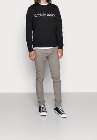 Černá mikina s bílým logem "Calvin Klein", béžové slim fit kalhoty a černé tenisky s bílou podešví. Čistý, moderní design.