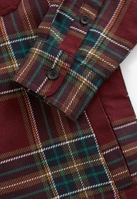 Gros plan sur un tissu à carreaux bordeaux montrant un poignet de chemise plié avec des boutons noirs et des lignes vertes, blanches, brunes et dorées dans le motif.