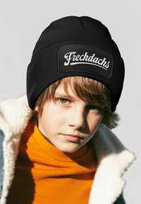 Black knit beanie with a textured surface and a prominent white logo patch reading "Frechdachs." Worn by a child with light brown hair.
