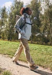 Giacca impermeabile azzurro chiaro con colletto alto, pantaloni beige e scarpe da trekking grigie con accenti turchesi, indossati mentre si cammina su un sentiero.