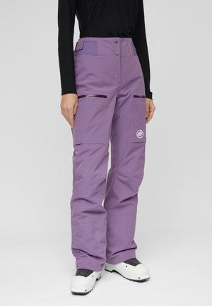 Personne portant un pantalon de neige violet taille haute avec poches zippées et des bottes de ski blanches, debout devant un fond uni.
