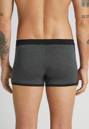 Der Rücken und die Beine eines Mannes sind sichtbar, der grau und schwarz gestreifte Boxer-Shorts trägt, mit Tätowierungen an beiden Armen vor einem schlichten Hintergrund.
