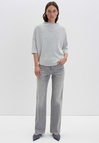 Hellgrauer, übergroßer Pullover mit hohem Halsausschnitt, kombiniert mit hellgrauen, geraden Jeans. Das Model steht selbstbewusst mit einer Hand auf der Hüfte.