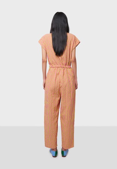 Mono de cuadros en rosa y amarillo con mangas cortas, cintura ceñida y piernas anchas. El cabello de la mujer cae por su espalda y se pueden ver unos zapatos coloridos.