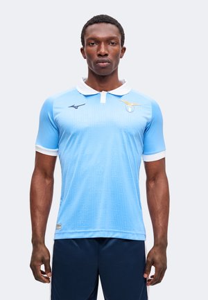 Homme portant un maillot de football bleu clair à manches courtes avec un col blanc et des bordures de manches blanches, debout devant un fond uni.