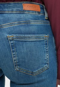 Jeans in denim blu con una vista ravvicinata della tasca posteriore, caratterizzata da cuciture arancioni e un'etichetta in pelle marrone con la scritta "ONLY". Texture liscia.