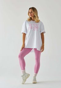 Biała, oversize'owa koszulka z różowym graficznym napisem, w zestawie z dopasowanymi różowymi legginsami i białymi trampkami z różowymi i beżowymi akcentami.