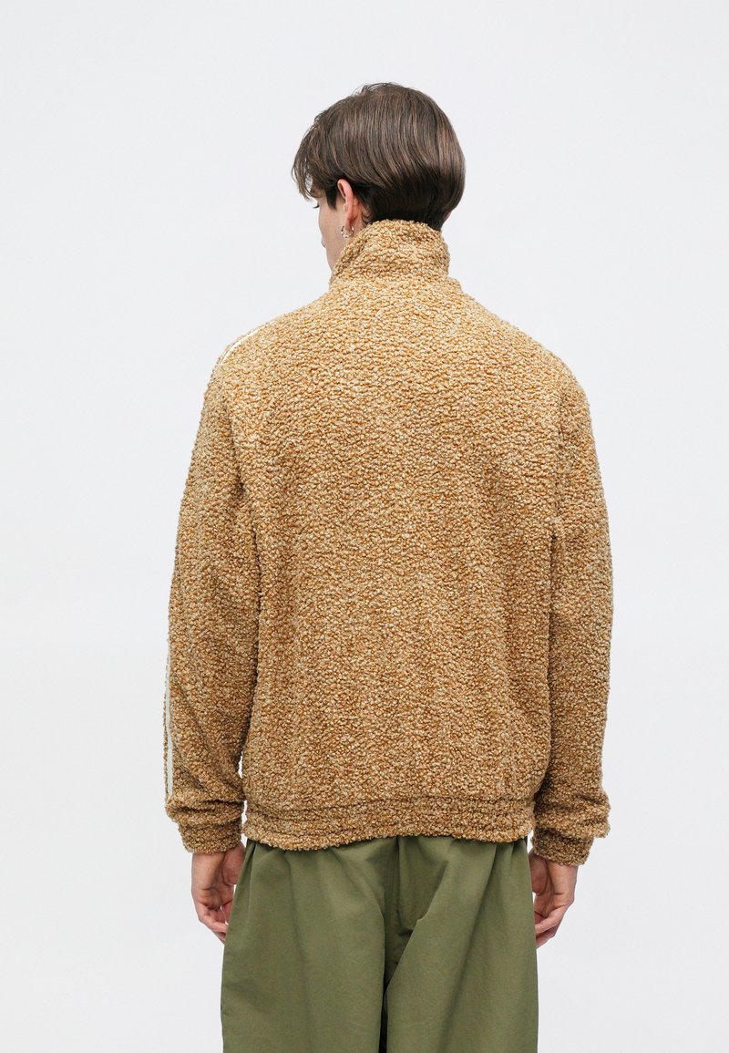 Brown textured fleece jacket with a high collar, featuring a relaxed fit and elastic cuffs, paired with olive green pants.