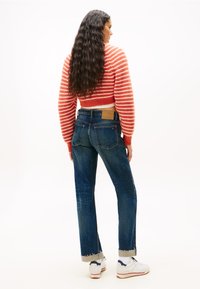 Kék farmernadrág, foszlott hemekkel és két hátsó zsebbel, amely egy vágott piros-fehér csíkos pulóverrel és fehér sportcipőkkel van párosítva.