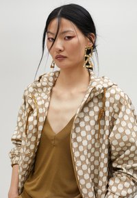 Brązowa i kremowa kurtka zapinana na zamek z kapturem, w połączeniu z brązową bluzką o głębokim dekolcie w kształcie V oraz dużymi złotymi geometrycznymi kolczykami.