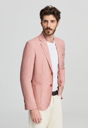 Blazer di lino rosa chiaro con una finitura testurizzata, rever a punta, due bottoni e tasche frontali, indossato su una t-shirt bianca e pantaloni color crema.
