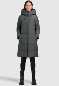 Long manteau gris duveteux avec capuche, doté d'une fermeture éclair à l'avant, de poches latérales et de cordons élastiques au col. Texture lisse et design épuré.
