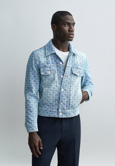 Homme portant une veste en denim à carreaux bleu clair sur une chemise blanche et un pantalon sombre, debout avec une main dans la poche, regardant sur le côté.