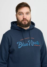 Tengerészkék kapucnis pulóver, puha anyagból készült, kenguruzsebb és húzózsinóros kapucni jellemzi. A "Blue Roots" logó kontrasztos kék és narancssárga nyomattal van ellátva.
