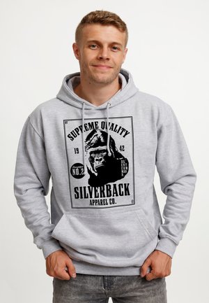 Młody mężczyzna z krótkimi włosami, ubrany w jasnoszary kaptur z czarną grafiką goryla i napisem "Silverback Apparel Co.".