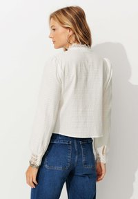 Blouse blanche texturée à manches longues, avec des accents en dentelle aux poignets et un design court. Portée avec un jean en denim foncé, vue de dos.