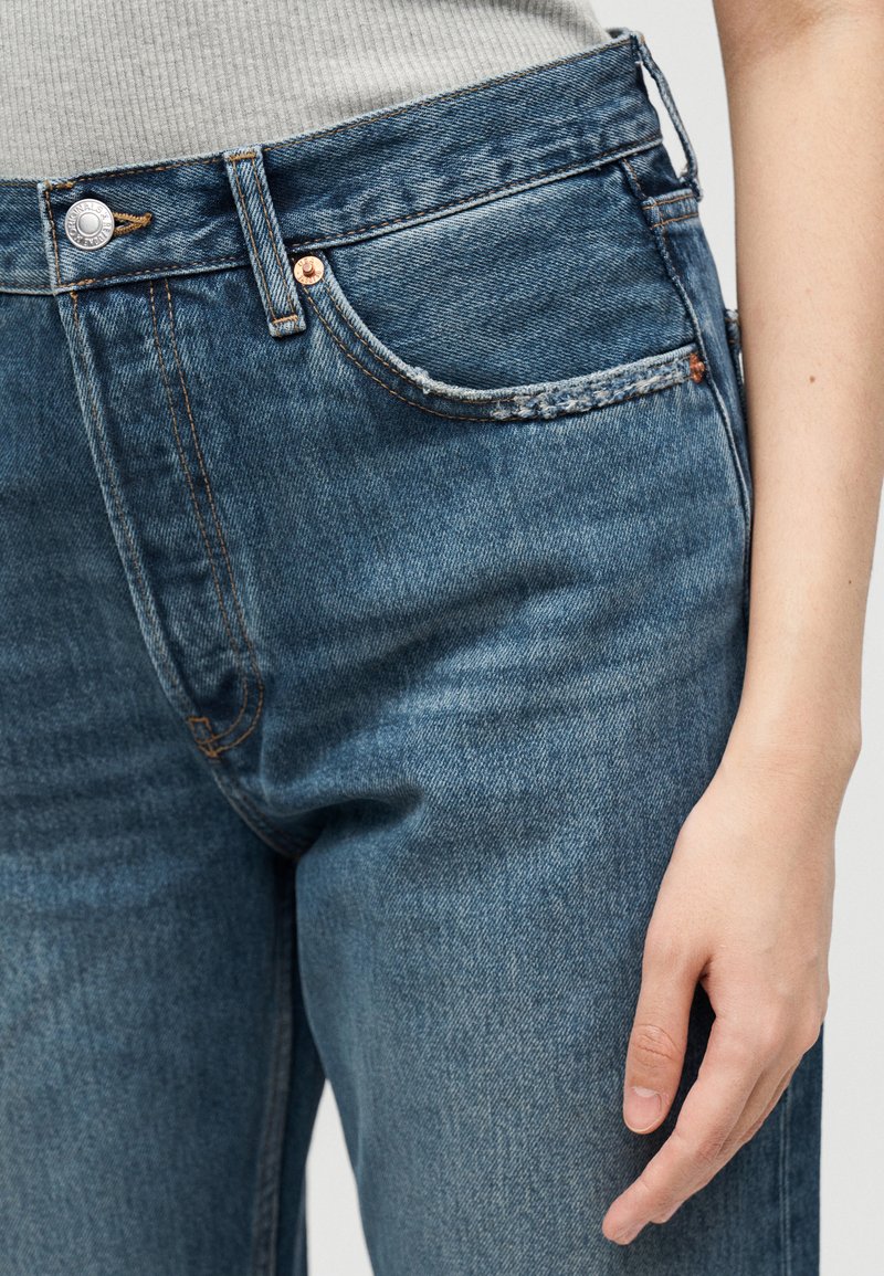 Nahaufnahme von mittelblauen Denim-Jeans mit Metallknopf und kleinem Vordertasche, getragen mit einem hellgrauen Ripptop.