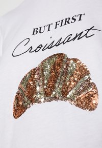 Vit t-shirt med svart texten "MEN FÖRST" ovanför en paillettdekorerad croissantdesign i nyanser av koppar, grön och guld. Slät textur.