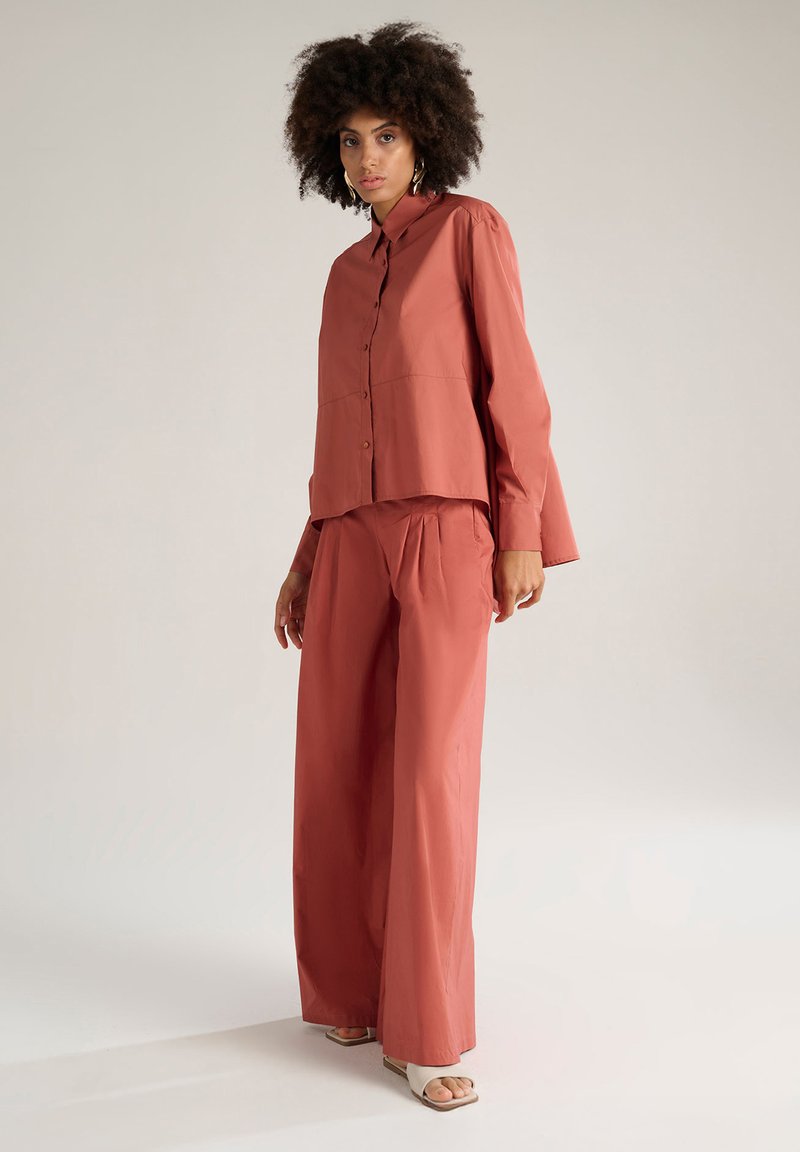 Camicia di colore ruggine, con una vestibilità ampia, maniche lunghe e bottoni, abbinata a pantaloni larghi coordinati. Tessuto liscio e design minimal.