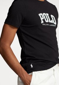Muž v černém tričku Polo Ralph Lauren s bílým logem na hrudi a malým vyšitým logem koně blízko lemu, v kombinaci s bílými kalhotami.