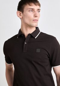 Brown polo shirt with white accents, short sleeves, a buttoned collar, and a logo patch on the chest, made from a soft fabric.
