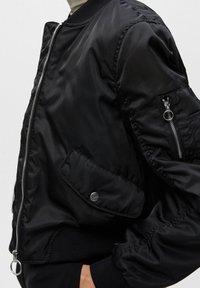 Chaqueta bomber negra con un exterior de nylon brillante, puños acanalados, cierre frontal con cremallera y múltiples bolsillos, incluido un bolsillo en el brazo con cremallera.