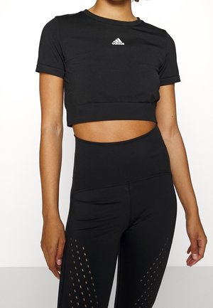 Femme portant un crop top noir Adidas à manches courtes et des leggings noirs taille haute avec des détails perforés sur les cuisses.