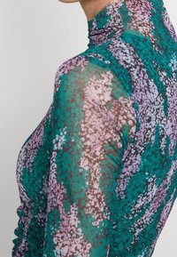 Gros plan d'une personne portant un haut transparent à col montant avec des motifs floraux abstraits en turquoise, violet et noir, et des manches longues.