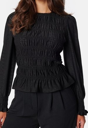 Kobieta nosząca dopasowaną czarną bluzkę z długim rękawem z marszczonym smockingiem, subtelną teksturą i falbanistym dołem, w połączeniu z czarnymi spodniami.