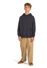 Sudadera gris oscuro con bolsillo frontal y pequeño logo, combinada con pantalones de pierna ancha color marrón claro y zapatillas marrones. La textura parece suave y relajada.