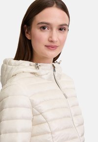 Lichte pufferjack in crèmekleur, met een rits aan de voorkant, kap met trekkoorden en een gequilt ontwerp met horizontale stiksels.