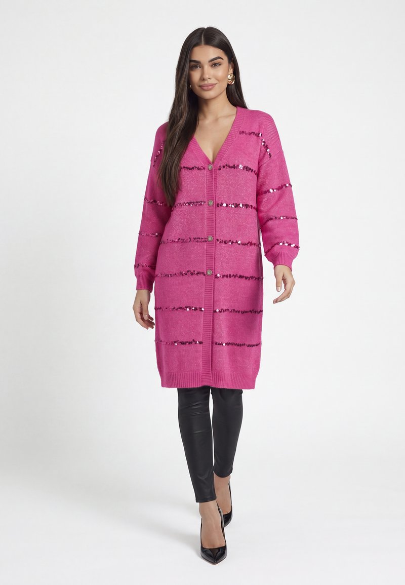 Femme portant un long cardigan rose avec des rayures horizontales en sequins, des leggings noirs et des talons hauts noirs, debout sur un fond blanc.