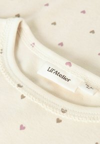 Bawełniany materiał w kremowym kolorze z małymi serduszkami w odcieniach różowym i brązowym, delikatny dekolt z falbanką oraz metka z napisem "Lil’Atelier."