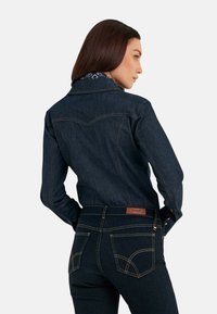 Camisa de denim oscuro con cuello clásico, costuras en contraste y canesú de estilo vaquero. Combinada con vaqueros oscuros que cuentan con bolsillos traseros y parche con logo.