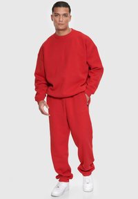 Roter Sweatshirt und passende Sweatpants aus weichem Stoff. Das Oberteil hat einen Rundhalsausschnitt und eine lockere Passform, mit minimalem Branding. Weiße Sneakers.