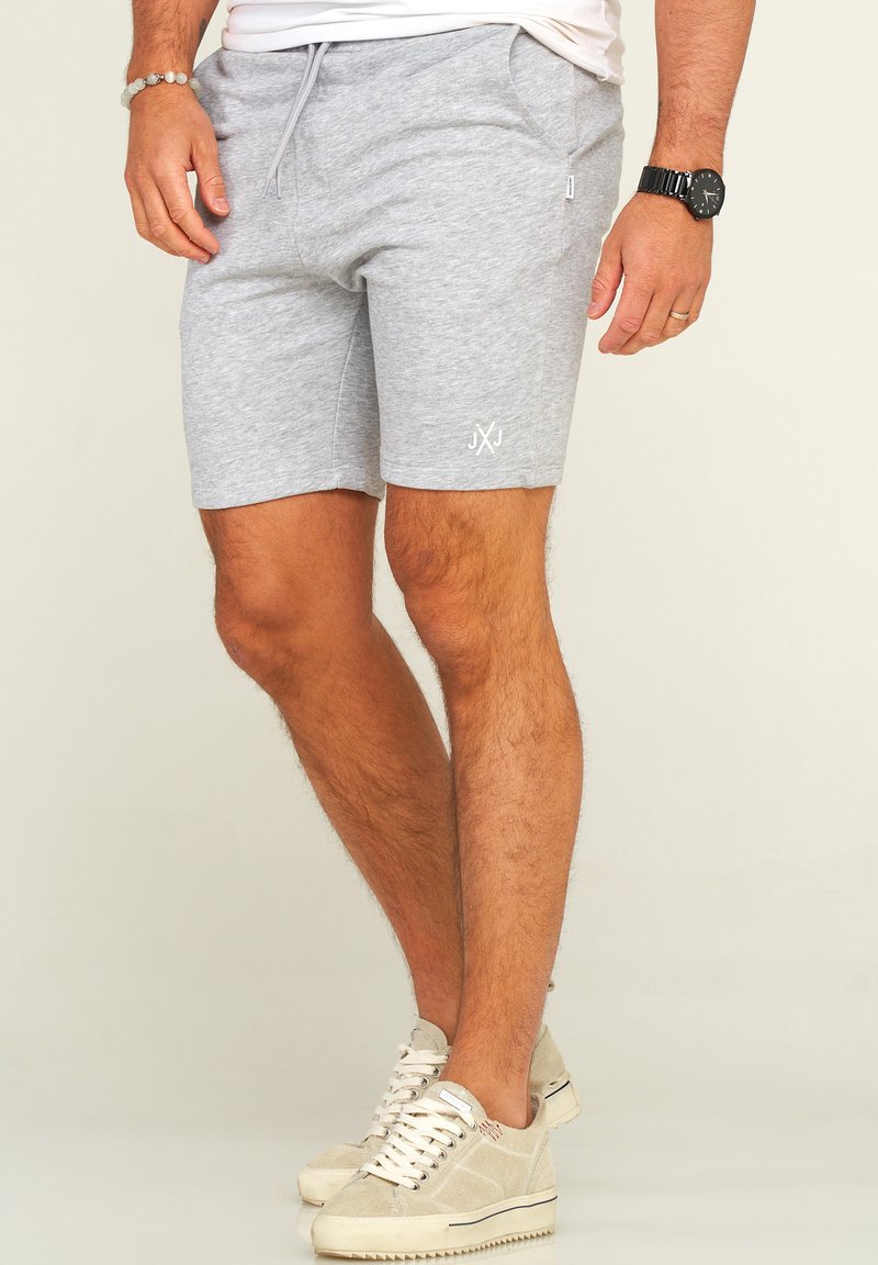 Graue Baumwoll-Shorts mit Seitentaschen, verstellbarem Bund und weißem gesticktem Logo am Bein. Getragen mit hellbeigen Sneakers.