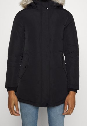 Manteau d'hiver noir avec capuche bordée de fourrure, poches avant à rabats, porté par une personne en jean bleu clair, sur fond uni.