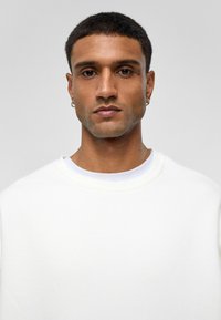 Witte sweatshirt van zacht materiaal, met een ronde halslijn en een oversized pasvorm, gedragen over een lichtblauwe shirt.