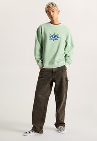Felpa verde chiaro con design grafico a stella, abbinata a jeans larghi marrone scuro e scarpe slip-on a scacchi bianco e nero.