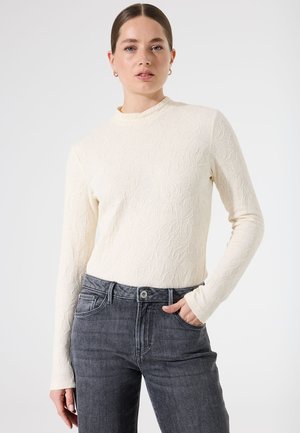 T-shirt à manches longues crème avec une texture florale complexe, col montant, associé à un jean gris foncé. Le tissu semble doux et ajusté.