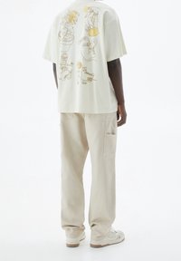 T-shirt oversize beige clair avec des illustrations de style cartoon en marron et jaune au dos, associé à un pantalon crème et des baskets.