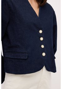 Blazer de mezclilla azul marino con escote en V, cuatro botones similares a perlas y un bolsillo en el pecho; diseño estructurado con un dobladillo ligeramente corto.