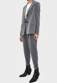 Kvinna iklädd en grå skräddarsydd kavaj och matchande högmidjade tapered byxor med ett svart bälte och svarta spetsiga ankelstövlar.