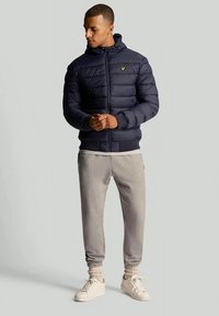 Homem em um casaco acolchoado azul-marinho com capuz, calças de moletom cinza claro, meias creme e tênis brancos, em pé contra um fundo simples.