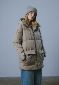 Kurtka puchowa w jasnym beżu z kapturem, przednim zamkiem błyskawicznym i dwiema dużymi kieszeniami. Połączona z niebieskimi dżinsami i szarym wełnianym czapkiem.