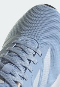 Gros plan d'une chaussure de course en mesh bleu clair avec tissu texturé, lacets assortis et vue partielle des bandes latérales blanches.