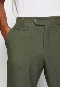 Olivgröna byxor i strukturerad tyg, med knappstängning, sidoficka och veckad frontdesign.