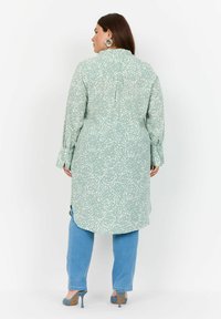 Lungo vestito verde menta con un motivo astratto bianco, polsini abbottonati e schiena parzialmente scoperta, abbinato a jeans azzurri e tacchi.