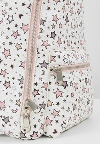 Sac à dos blanc avec une finition texturée, arborant des motifs d'étoiles et de lunes multicolores. Comprend une poche avant zippée avec des ferrures argentées.