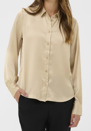 Kvinde iført en beige langærmet knappet skjorte med krave og sorte bukser, med den ene hånd i lommen mod en ensfarvet baggrund.